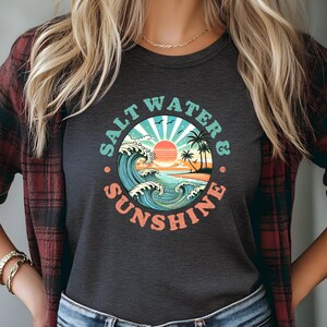 May include: A dark gray t-shirt with a vintage-style graphic of a sunset over the ocean with waves crashing. The graphic is surrounded by the text "Salt Water & Sunshine".