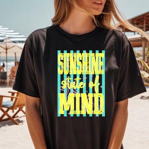 K&ouml;nnte beinhalten: Schwarzes T-Shirt mit dem Text "SUNSHINE state of MIND" in Gelb und Hellblau. Der Text befindet sich in einem hellblau-gelb gestreiften Rechteck. Das T-Shirt wird am Strand getragen.