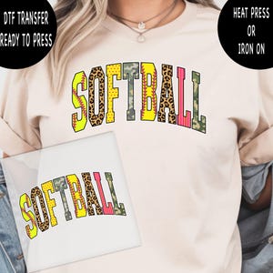 May include: A cream-colored t-shirt featuring the word "SOFTBALL" in a bold, multi-patterned design. The letters incorporate leopard print, camouflage, and pink stripes. The image also shows a DTF transfer ready to press.