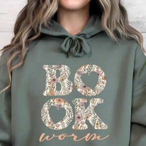 May include: Sage green hoodie with the word "BOOK" and "OK" in large floral letters, with a heart shape in the "O"s. The word "worm" is written in a cursive font below. The hoodie has a drawstring and a front pocket.