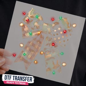May include: A decorative bow transfer featuring a mix of plaid and polka dot patterns in light tan and gold tones. The bow is adorned with a string of small, glowing Christmas lights in red, green, and warm white colors. Text reads DTF TRANSFER.