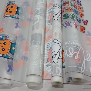 May include: Four rolls of clear vinyl with different designs printed on them. The designs include a pumpkin with a syringe, a ghost, a hand with a basketball, and a colorful alphabet.