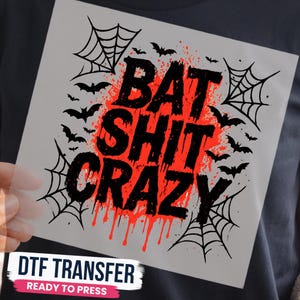 May include: A square DTF transfer sheet featuring the black text BAT SHIT CRAZY layered over a red paint splatter effect. The design includes black spider webs in the corners and multiple black bat silhouettes scattered throughout the graphic.