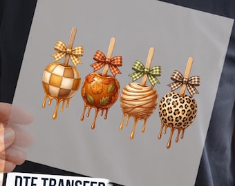 May include: A DTF transfer featuring four caramel apples on sticks, each with a unique pattern: a tan and white checkered design, an orange leaf print, a white drizzle, and a brown leopard print, all topped with matching fabric bows.