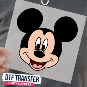 May include: A DTF transfer sheet featuring a graphic of Mickey Mouse's face with black ears, a peach-colored face, and a smiling mouth. The text at the bottom reads DTF TRANSFER READY TO PRESS in bold black and white letters on a pink background.
