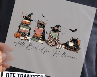 May include: A dark gray t-shirt featuring a graphic print of four black cats reading books. The design includes pumpkins, a skull, bats, and the text "All Booked for Halloween" in a cursive script. The cats wear witch hats and floral crowns.