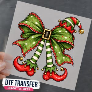 May include: A DTF transfer featuring a green bow with white polka dots and red scalloped edges, centered with a gold buckle. Two striped green and white elf legs extend downward, ending in curled red shoes with gold jingle bells attached.