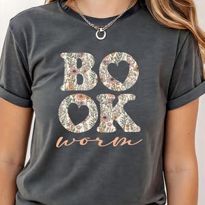 May include: Dark gray t-shirt with the word "BOOK" and "OK" in a floral pattern, with heart-shaped cutouts in the O's. The word "worm" is written in a cursive font below the other words. The shirt has short sleeves.