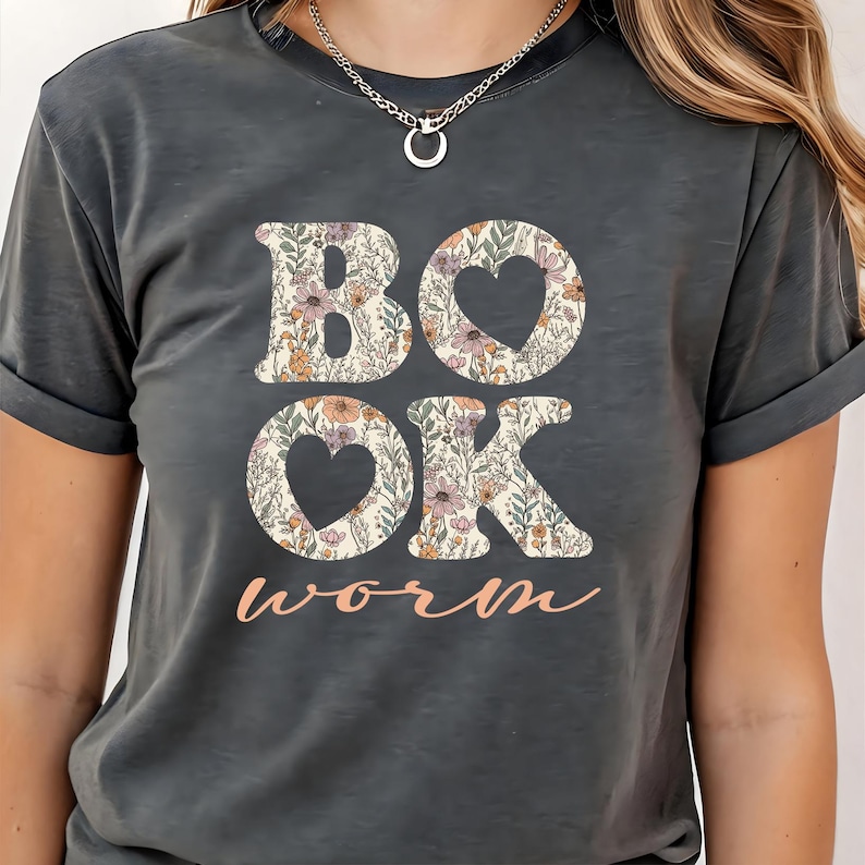 May include: Dark gray t-shirt with the word "BOOK" and "OK" in a floral pattern, with heart-shaped cutouts in the O's. The word "worm" is written in a cursive font below the other words. The shirt has short sleeves.