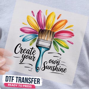 May include: A DTF transfer sheet featuring a paintbrush with colorful, petal-like paint strokes radiating outward in yellow, pink, orange, blue, and green. The black text reads Create your own Sunshine. The design is on a clear film sheet.