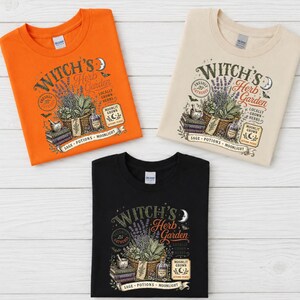 May include: Three crew neck t-shirts in orange, beige, and black. Each features a graphic print titled Witch's Herb Garden with illustrations of a basket of herbs, books, a mortar and pestle, a potion bottle, a crescent moon, and a bat.