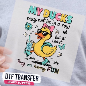 May include: A DTF transfer featuring a yellow duck wearing sunglasses, a pink bow, and a backpack while roller skating. The duck holds a blue drink. Text reads: MY DUCKS may not be in a row But at Least They are having FUN. Includes flowers and butterflies.