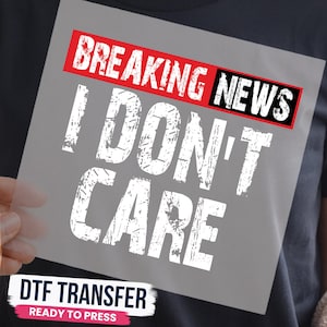 May include: A DTF transfer sheet featuring a graphic design with the text BREAKING NEWS in a red and black box, followed by the words I DON'T CARE in large, distressed white lettering. The design is printed on a clear film for heat application.
