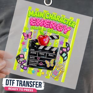 May include: A DTF transfer sheet featuring the text Main Character Energy Starts In Second Grade in neon green and pink checkered letters. The design includes a red apple, a black film clapperboard, film strips, butterflies, stars, and popcorn.