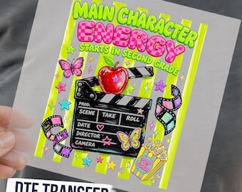 May include: A DTF transfer sheet featuring the text Main Character Energy Starts In Second Grade in neon green and pink checkered letters. The design includes a red apple, a black film clapperboard, film strips, butterflies, stars, and popcorn.