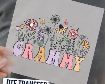 May include: A DTF transfer sheet featuring the word GRAMMY in a retro, bubbly, multi-colored font. Above the text is a botanical illustration of various flowers, including daisies, lavender, and dandelions, with small butterflies and stars.
