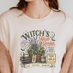 May include: A cream-colored t-shirt featuring a graphic print with the text "Witch's Herb Garden". The design includes a basket of lavender, a mortar and pestle, stacked books labeled "Herbology" and "Potions", a bottle, and a crescent moon.