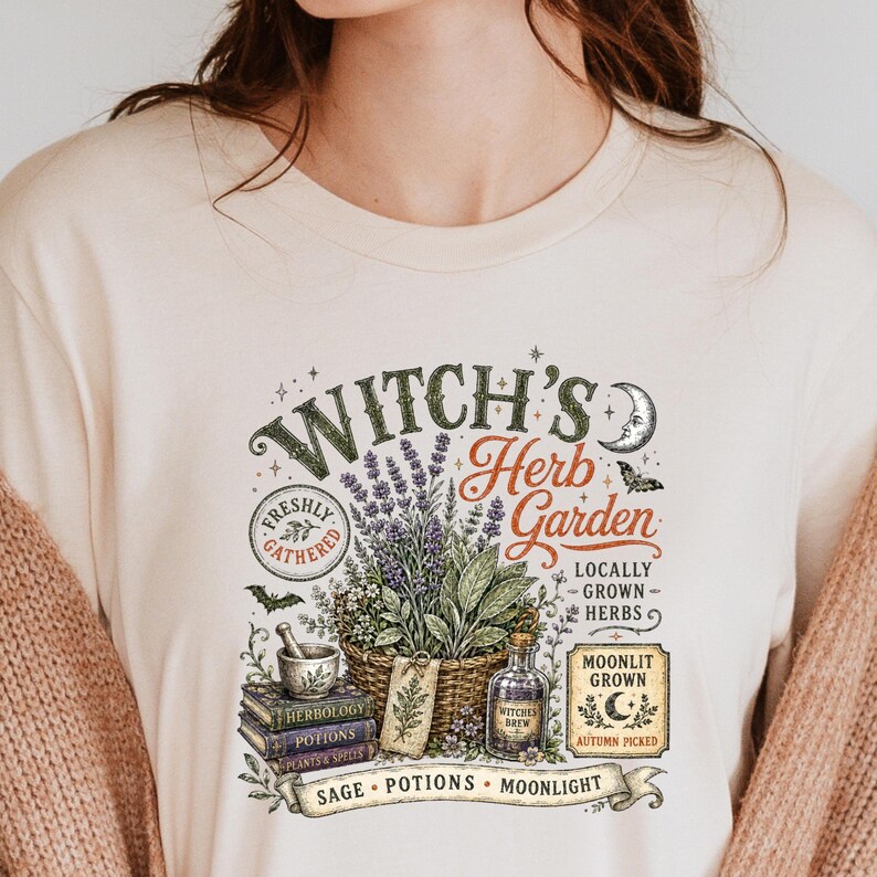 May include: A cream-colored t-shirt featuring a graphic print with the text "Witch's Herb Garden". The design includes a basket of lavender, a mortar and pestle, stacked books labeled "Herbology" and "Potions", a bottle, and a crescent moon.