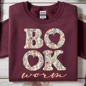 May include: Burgundy sweatshirt with the words "BOOK WORM" in a floral design. The letters "B" and "K" are filled with a floral pattern, and the "O"s are heart-shaped. The word "worm" is in a cursive font.