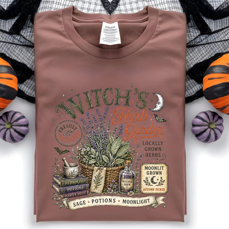 May include: A mauve crew neck t-shirt featuring a graphic print of a basket of herbs, a mortar and pestle, stacked books labeled Herbology, Potions, and Spells, and a bottle labeled Witches Brew, with text reading Witch's Herb Garden.