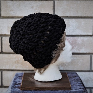 May include: A black knitted beanie hat with a chunky knit texture. The hat is on a mannequin head.