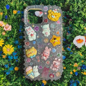 Può includere: Una custodia per telefono trasparente con uno sfondo scintillante e vari personaggi dei cartoni animati colorati, tra cui un orso, un gatto, un coniglio e una stella. La custodia è decorata con piccoli fiori e strass.