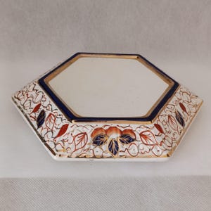 Könnte beinhalten: Ein sechseckiger Keramikuntersetzer mit einer glatten weißen Mitte, die von einem tiefblauen Rand mit Goldakzenten umgeben ist. Die abgeschrägten Kanten sind mit einem Muster aus orangefarbenen und blauen Blättern verziert.