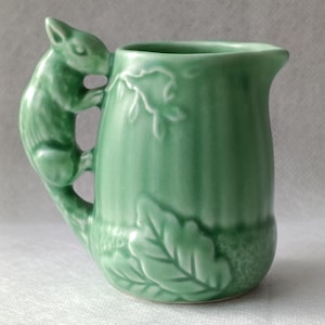 May include: A light green ceramic creamer featuring a sculpted squirrel handle. The body of the pitcher is decorated with embossed vertical lines and detailed oak leaf patterns at the base, finished with a glossy glaze and a small pouring spout.