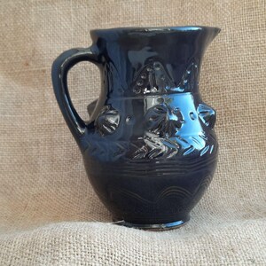 May include: A dark blue ceramic pitcher with a handle and decorative embossed floral and leaf patterns. The pitcher has a glossy finish and a wide opening. The height of the pitcher is approximately 8 inches.