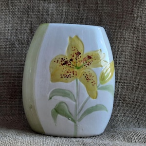 Könnte beinhalten: Weiße Keramikvase mit einer gelben Lilie, die auf die Vorderseite gemalt ist. Die Lilie hat braune Flecken und grüne Blätter.