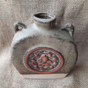 May include: A brown and tan ceramic vase with a circular design in red and white. The vase has two handles and a rounded body.