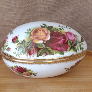 以下が含まれることがあります： 金色の縁取りが付いた、卵型の白い磁器製の小物入れ。黄色、ピンク、赤のバラと緑の葉のフローラルパターンで装飾されています。木製のトレイの上に置かれています。