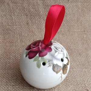 Puede incluir: Adorno esférico de cerámica blanca con un diseño floral de rosas burdeos y blancas, acentuado con hojas verdes. El adorno tiene tres pequeños agujeros y una cinta roja para colgar.