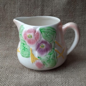 May include: A small, white ceramic pitcher with a pink handle and rim. The pitcher is decorated with pink, purple, and green floral designs. Yellow accents are also present. The pitcher has a spout for pouring.