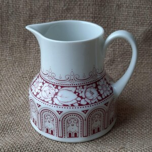 Può includere: Brocca in ceramica bianca con un motivo floreale e geometrico rosso. La brocca ha un beccuccio e un manico curvo. Il design include rose, foglie e forme ad arco. Un pezzo decorativo per servire bevande.