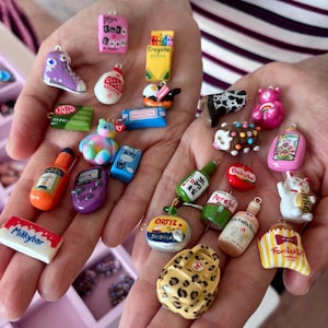 May include: A collection of miniature charms in various shapes and colors, including a purple sneaker, a pink book, a yellow crayon box, a green KitKat bar, a tie-dye bear, a hot sauce bottle, a purple game console, and a white Milkybar chocolate bar.
