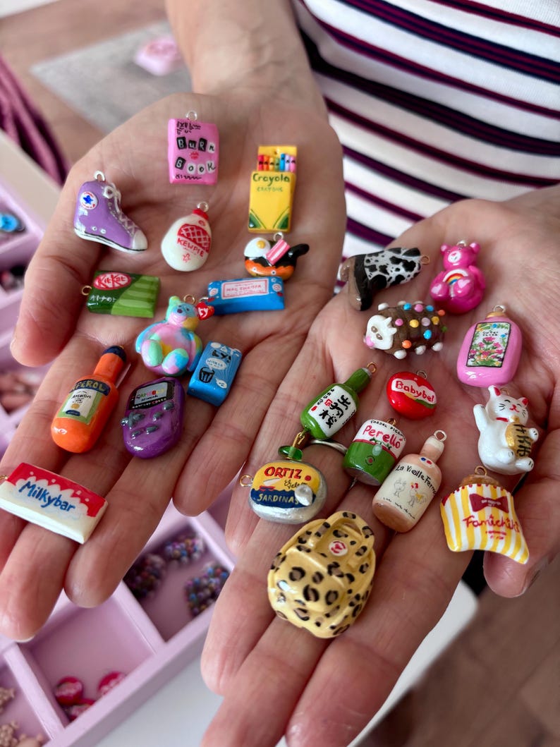 May include: A collection of miniature charms in various shapes and colors, including a purple sneaker, a pink book, a yellow crayon box, a green KitKat bar, a tie-dye bear, a hot sauce bottle, a purple game console, and a white Milkybar chocolate bar.