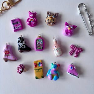 May include: A collection of small, colorful charms including a pink bear, a leopard-print creature, a purple Furby, a pink cowboy boot with a heart, a black and white cow-print boot, a pink bottle, a crayon box, a mushroom, and a pink sneaker.