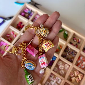 May include: A gold-toned metal keychain featuring multiple miniature charms including a leopard-print creature, a yellow and white striped snack bag, a pink strawberry milk carton, a pink KitKat bar, a sardine tin, and a green bottle with Japanese text.