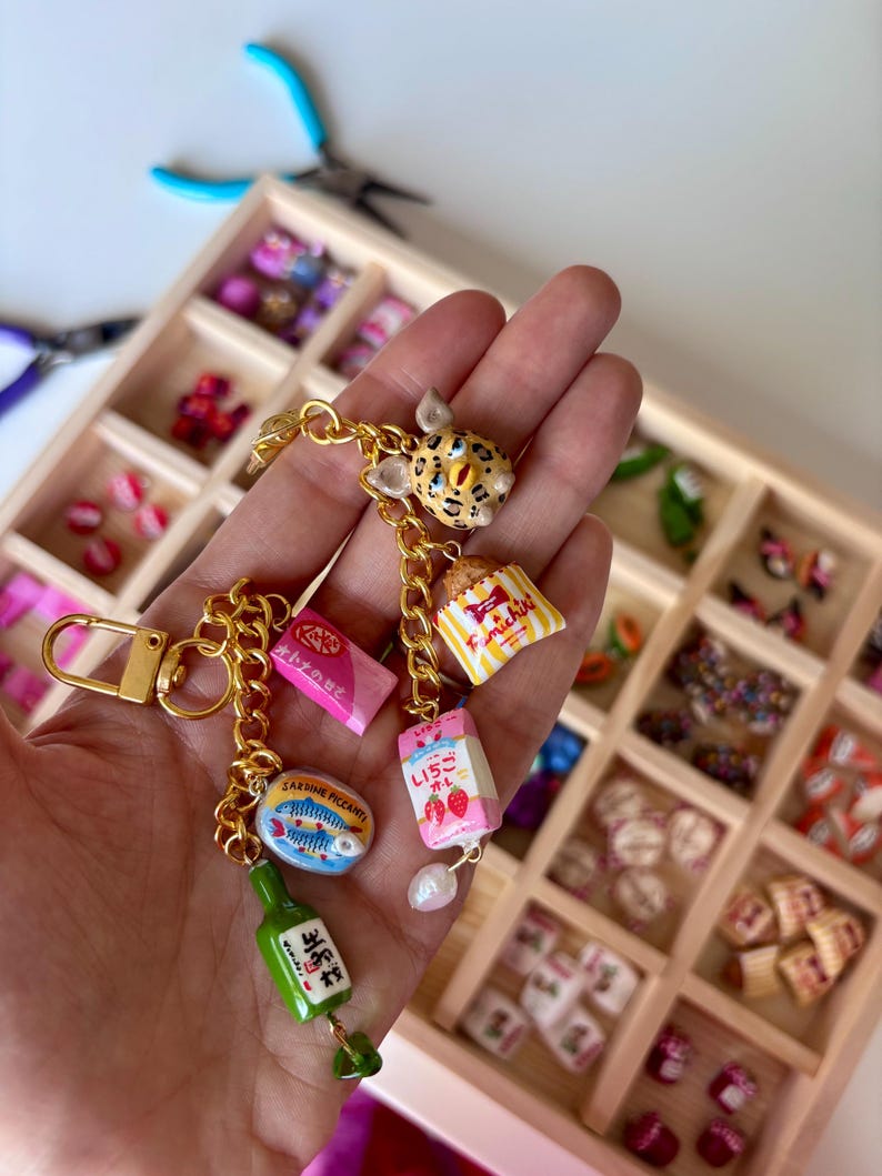 May include: A gold-toned metal keychain featuring multiple miniature charms including a leopard-print creature, a yellow and white striped snack bag, a pink strawberry milk carton, a pink KitKat bar, a sardine tin, and a green bottle with Japanese text.