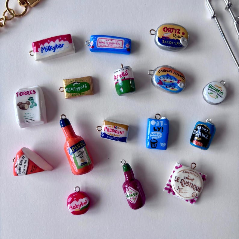 May include: A collection of miniature food and beverage charms featuring various brand labels including Milkybar, Kerrygold, Perell&oacute;, Ortiz, Sardine Piccante, Boursin, Torres, Pr&eacute;sident, Oatly, Heinz Beanz, Port Salut, Aperol, Babybel, Tabasco, and Le Rustique.