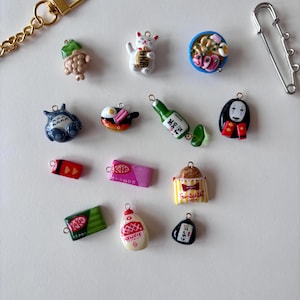 May include: A collection of miniature charms including a green taiyaki, a white maneki-neko, a bowl of ramen, a blue Totoro, a fried egg, a green bottle, a black spirit figure, a red candy, a pink KitKat, a striped bag, a green KitKat, a Kewpie bottle, and a black onigiri.