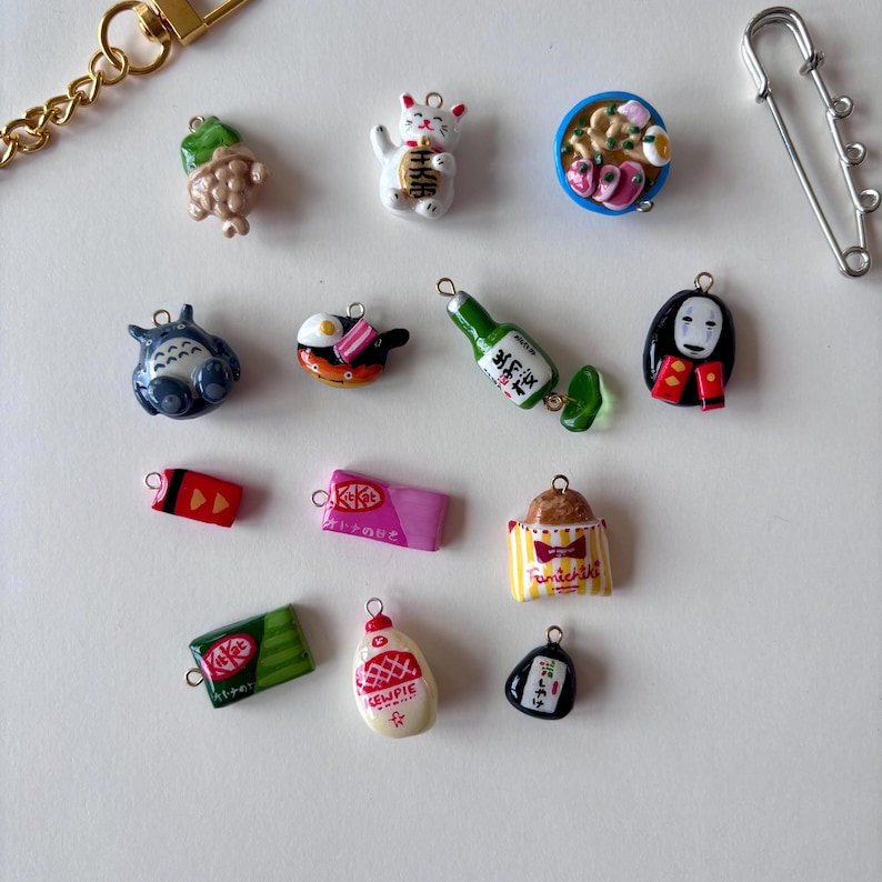 May include: A collection of miniature charms including a green taiyaki, a white maneki-neko, a bowl of ramen, a blue Totoro, a fried egg, a green bottle, a black spirit figure, a red candy, a pink KitKat, a striped bag, a green KitKat, a Kewpie bottle, and a black onigiri.