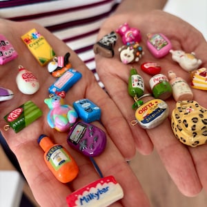 May include: A collection of miniature charms featuring various food, drink, and pop culture items including a yellow Crayola box, a green KitKat bar, an orange Aperol bottle, a Milkybar, a purple handheld game console, and an Ortiz sardine tin.