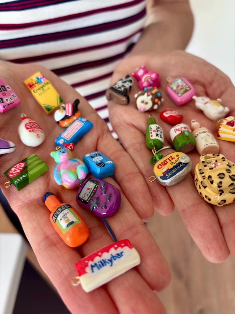 May include: A collection of miniature charms featuring various food, drink, and pop culture items including a yellow Crayola box, a green KitKat bar, an orange Aperol bottle, a Milkybar, a purple handheld game console, and an Ortiz sardine tin.