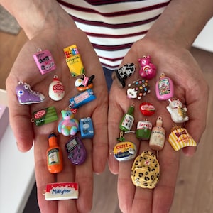 May include: A collection of miniature charms featuring various designs including a pink Burn Book, yellow Crayola crayons, a purple sneaker, a KitKat bar, a colorful bear, an Aperol bottle, a Milkybar, a cow print boot, a hedgehog, and a leopard backpack.