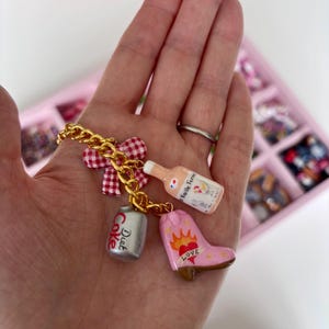 May include: A gold-tone chain charm bracelet featuring four distinct charms: a red and white gingham bow, a miniature bottle labeled La Vieille Ferme, a silver-colored Diet Coke can, and a pink cowboy boot with a heart and flame graphic.