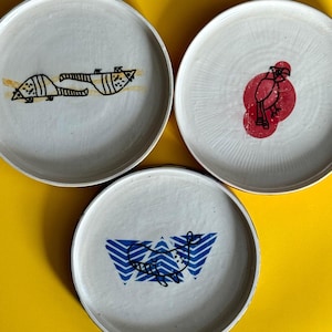 May include: Three round ceramic plates with a white finish. The top left plate features a black line drawing of an armadillo. The top right plate displays a black line drawing of a bird over a red circle. The bottom plate shows a blue whale graphic.