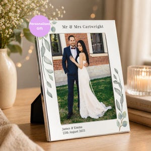 May include: A silver photo frame displaying a wedding photograph of a couple. The frame features a white border adorned with green leaf designs. The top of the frame reads "Mr & Mrs Cartwright" and the bottom reads "James & Emma 25th August 2023".