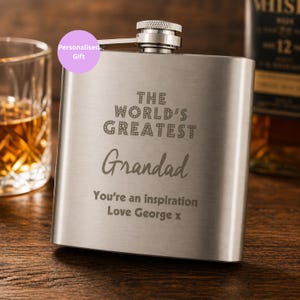 May include: A silver-coloured hip flask engraved with "THE WORLD'S GREATEST Grandad". The text "You're an inspiration Love George x" is also present. A glass of amber liquid and a bottle of whiskey are in the background.